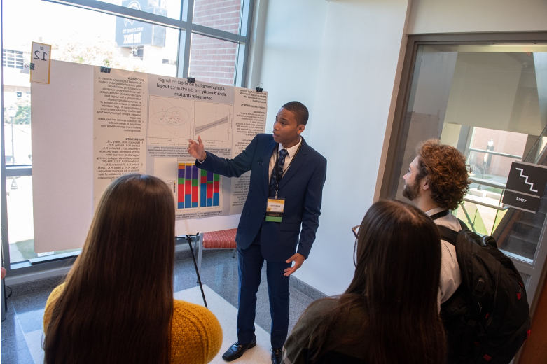 Forensics (Biological Sciences) major Jordan Cole received a 2022 Drapeau Summer Research Grant to study the effects of algae and fungi on wood decay under the mentorship of Biological Sciences professor, Dr. Kevin Kuehn. Here he is shown explaining his DCUR-funded research project to UGS 2023 attendees. 